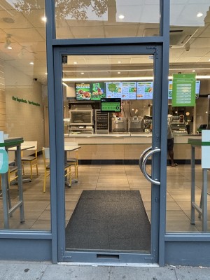 Glass door with grey frame. Door handle to the right, push to open. Glass walls on either side