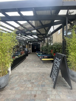 From streer facing venue. Opening ahead. A sign frame on right. Outdoor seating beyond it. Path down centre to doors at back.