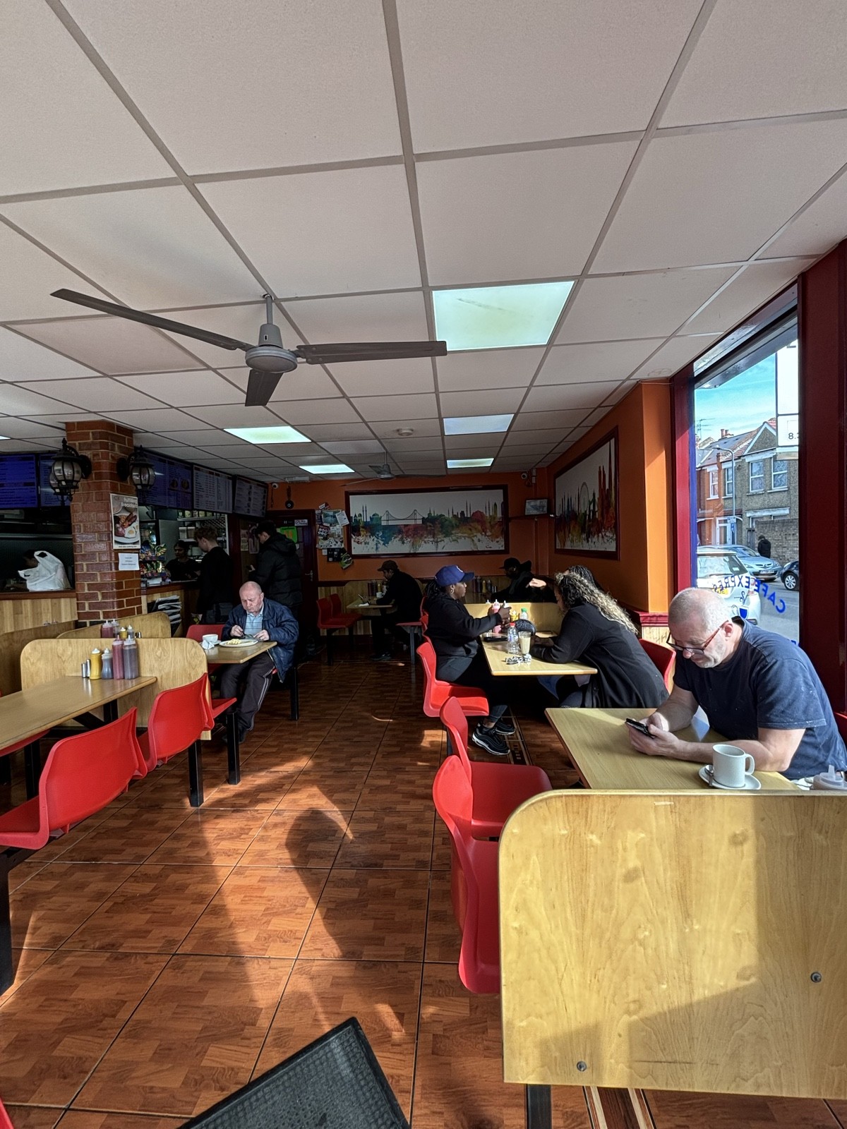 From entrance facing right, tables and chairs along right glass wall, tables and chairs along wall at the back. Large paintings on orange walls