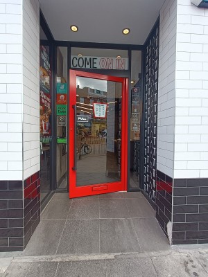 From street, facing venue. Opening ahead. Glass door with red, metal frame at back.