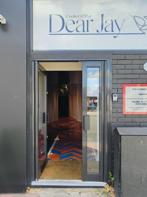 From sidewalk facing venue. Grey framed preceding glass door ahead. Push grey handle in centre to open.