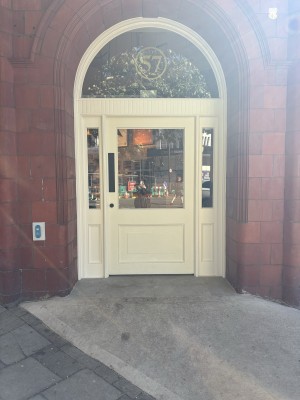 From sidewalk facing straight, white wooden door with glass windows with arch above. Request for assistance button to the left of door.
