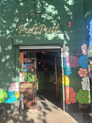 From sidewalk facing venue. Brown framed glass door ahead. Push golden handle in centre to open.
