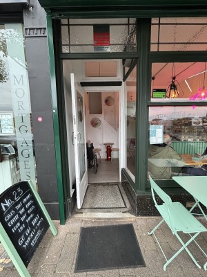 From sidewalk facing venue, green archway preceding doorway. Door with white doorframe and glass centre. Door handle to the right, pull to open.