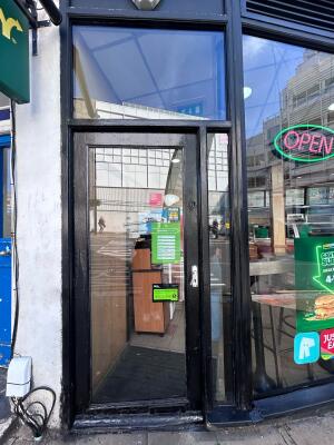 From sidewalk facing straight. Glass wall with black metal frames ahead. Push lever on right to open.