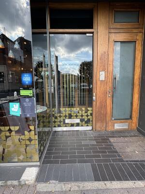 Glass door with metal handle on left hand side, pull to open. Black tiling preceding entrance