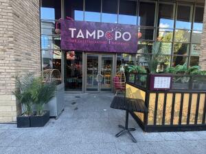From sidewalk facing venue entrance. Opening ahead leading to main doors of venue and outdoor seating on both sides.