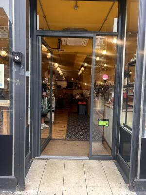 From sidewalk facing entrance. Black framed archway ahead. Glass door with black metal frame. Push metal handle on right to open.