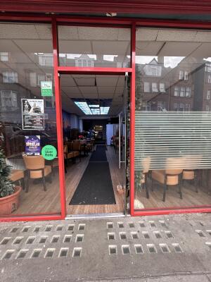 From sidewalk facing venue. A slope up towards door. Glass door with red metal frame ahead. Push metal handle on left to open.