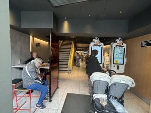 From entrance facing straight. High table and stools on left. Staircase behind. Kiosks on right. Path in centre leading to more seating at back.