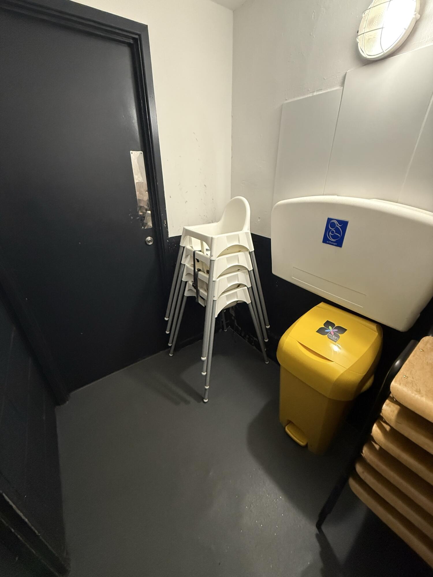 From inside bathroom facing corner. Baby high chairs stacked left of baby changing station. Yellow bin below station on right.