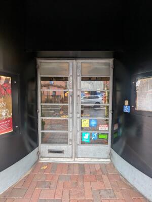 Grey frame glass doors. Metal handles in center. Pull to open. Doorbell for assistance on its right.