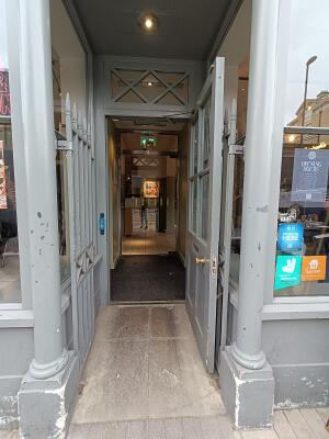 From street, facing venue. Open archway ahead to glass door with grey wooden frame ahead. Pull from left to open. Doorbell for assistance on left.