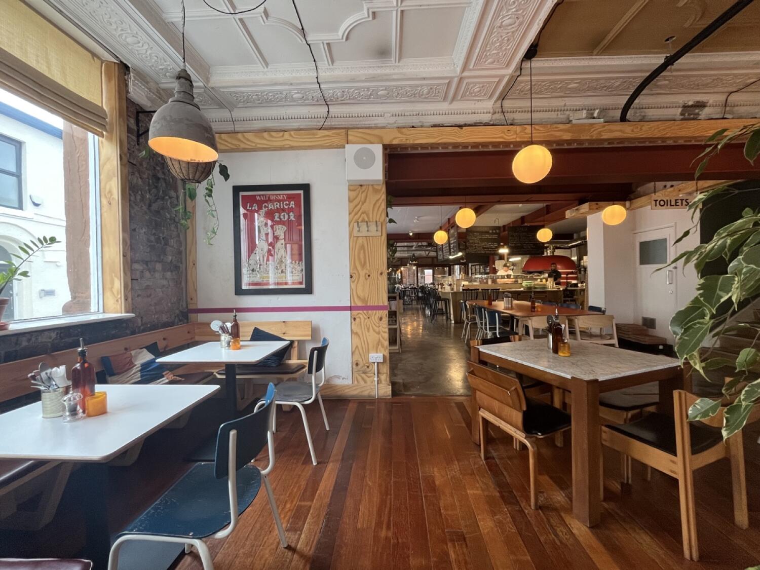 From door facing left. Half booth, tables and chairs along left. Path in centre. Archway to open kitchen at back. Table and chairs on right.
