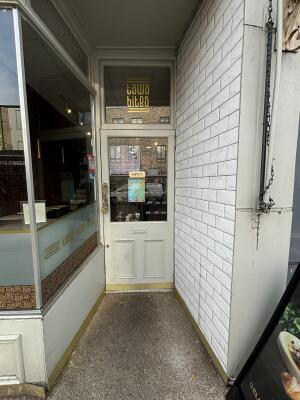 From sidewalk facing venue. Off white door with golden handle on left ahead. Pull door to open.