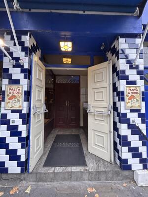 From sidewalk facing venue.
Blue wooden doors with blue frames ahead. Pull from center to open. Second set of doors at back.