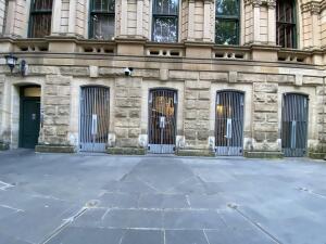 From street facing building. Green door far left. Path in centre to metal gates span center to right, each with vertical metal bars and handles.