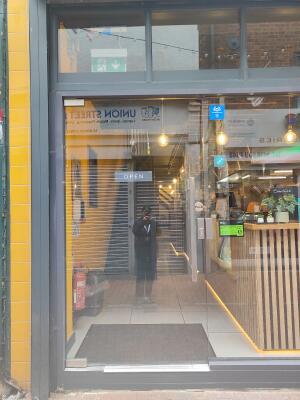 From sidewalk facing venue. Grey framed glass door ahead. Push silver bar on left to open.