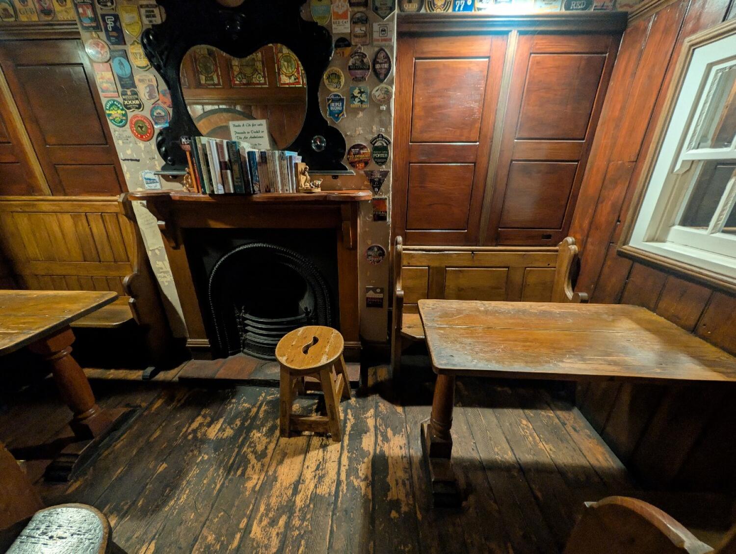 From wooden archway facing straight. Tables and benches on both sides. Fireplace at back center. Stool in front.