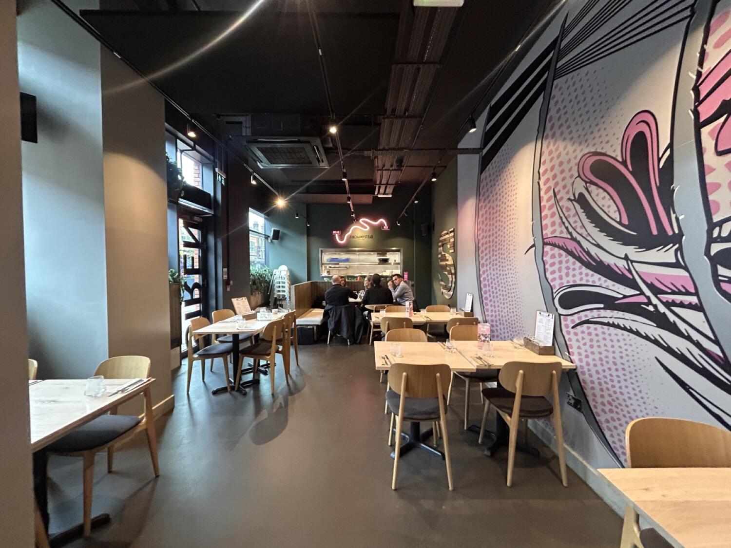 From pillar facing straight. Chairs on left. Second wall partition behind. Chairs behind. Chairs on right. Half-booth at back. Kitchen behind.