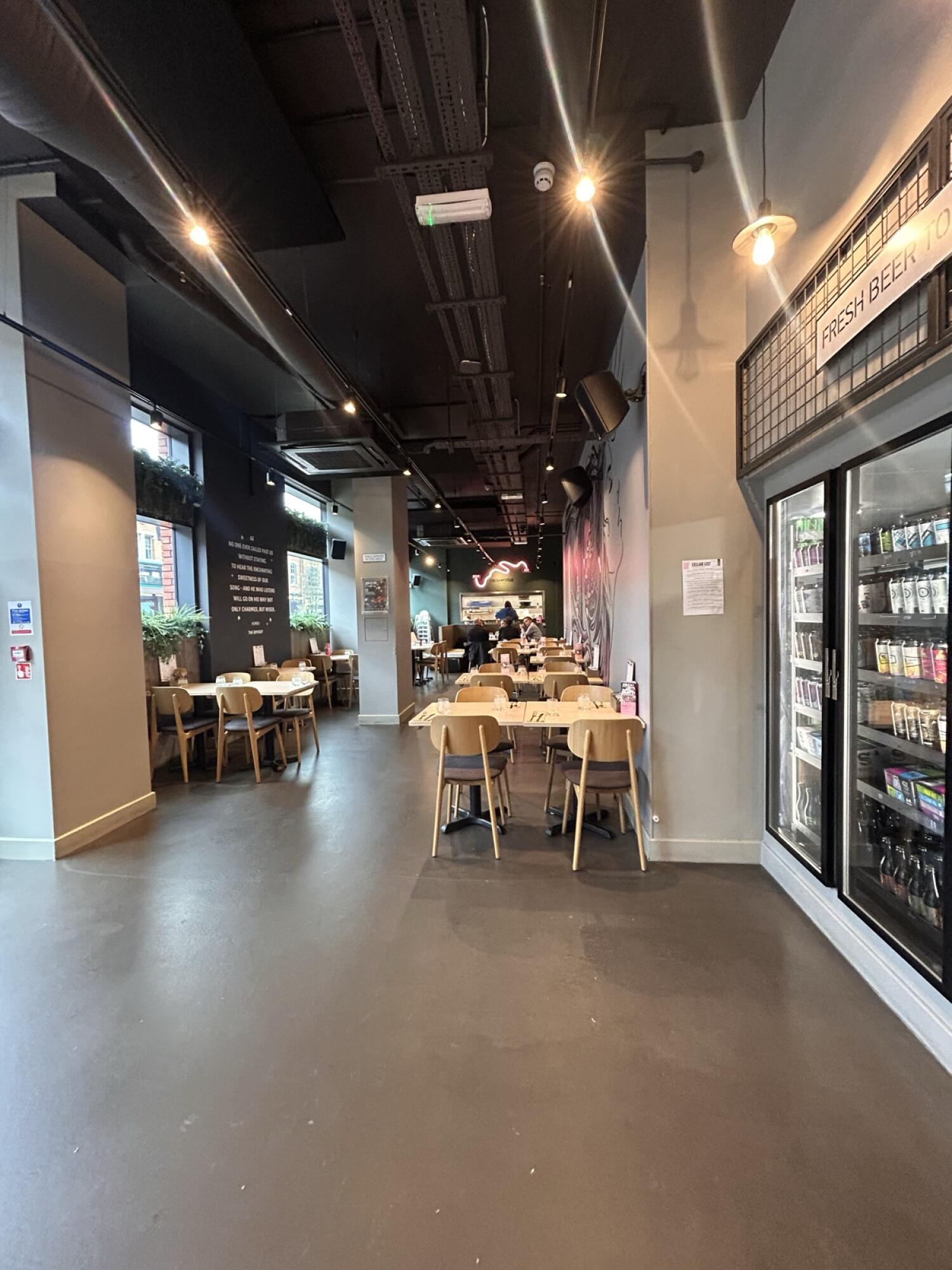 From after shelf facing straight. Wall partition on left. Chairs behind. Pillar at back. Fridge on right. Chairs and tables behind.