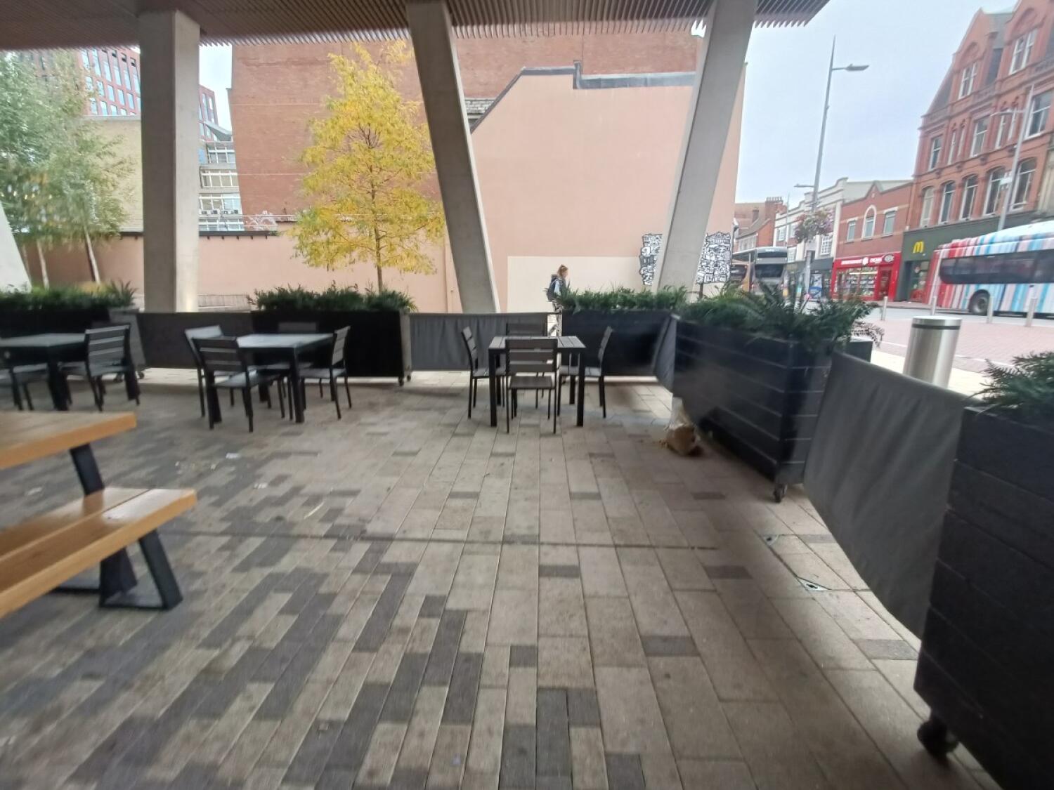 From doorway, facing straight. Planters and restaurant barriers along right and back. Tables and benches on left. Tables and chairs along back.