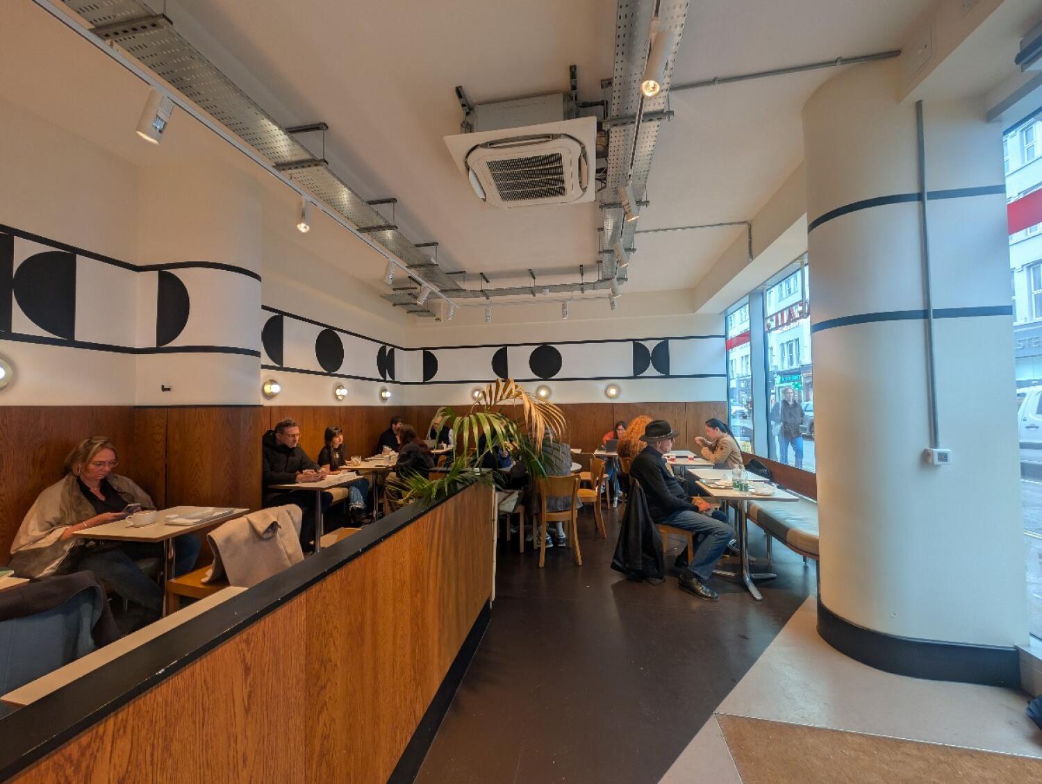 From entrance facing right. Tables, chairs, and half booths along both sides and back. Wooden barrier on along left middle. Tables and chairs behind.