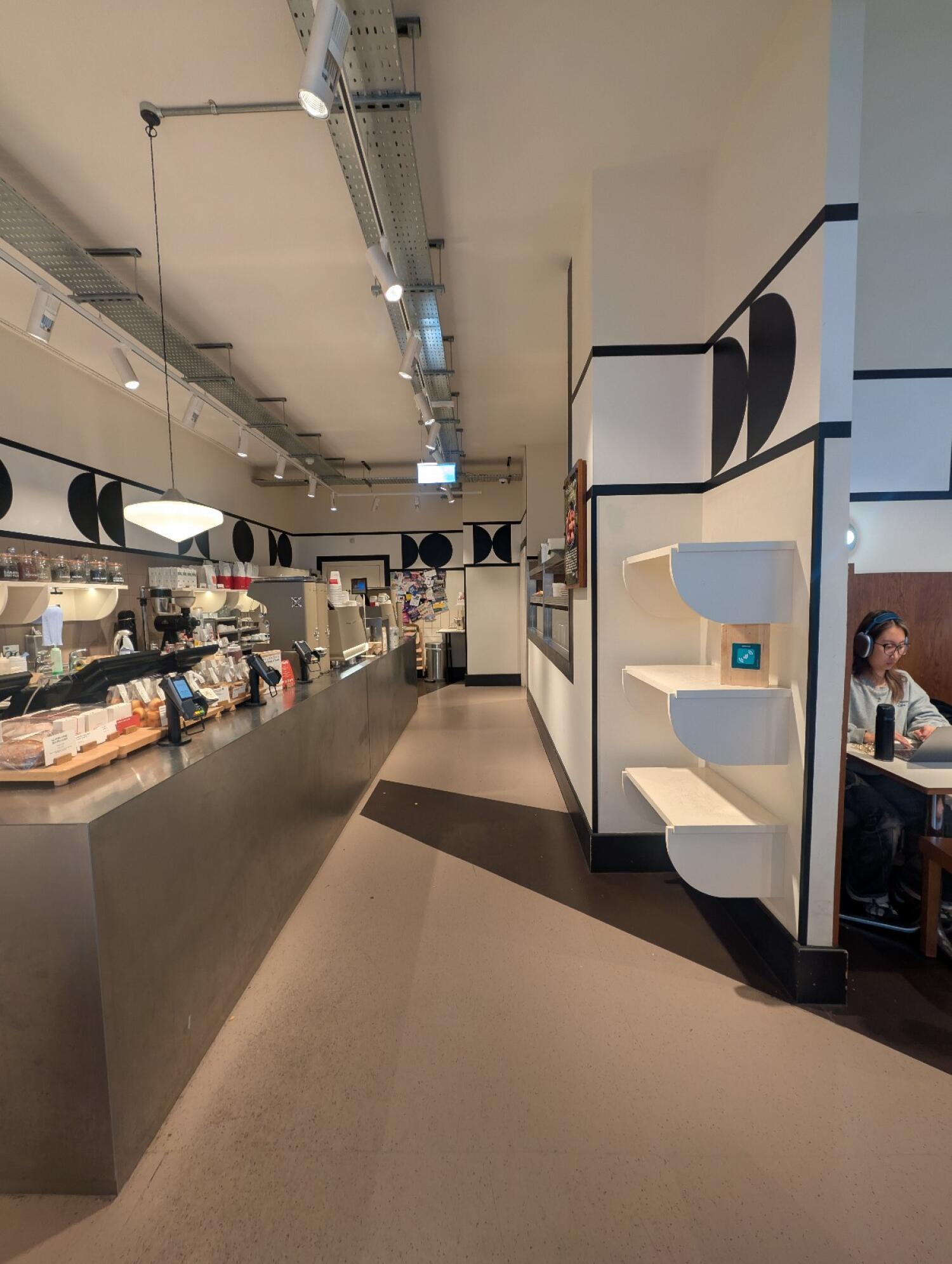 From left of wooden barrier facing straight. Bakery counter along left. Path in center to bathroom. Open kitchen along right.