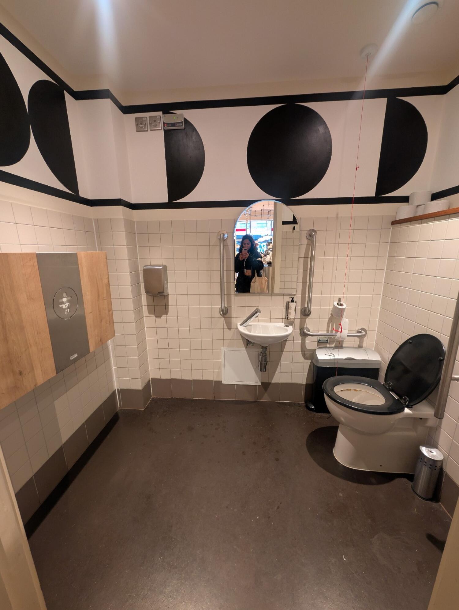 From bathroom door facing straight. Toilet seat on right wall. Pull cord behind. Sink on back right. Baby change table on left wall.