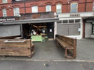From street, facing venue. Wooden fence on left. Opening ahead. Bench on right.