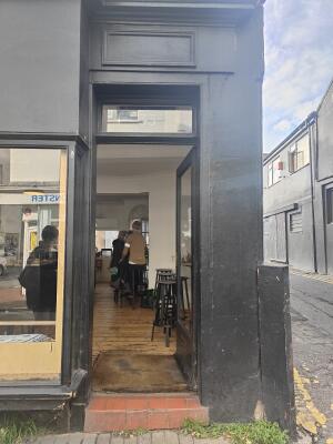 From sidewalk facing straight. Black framed glass door ahead. Push golden handle on left to open.