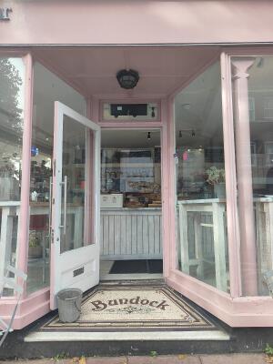 From sidewalk facing venue. Pink archway ahead. White door at back. Pull silver handle on right to open.