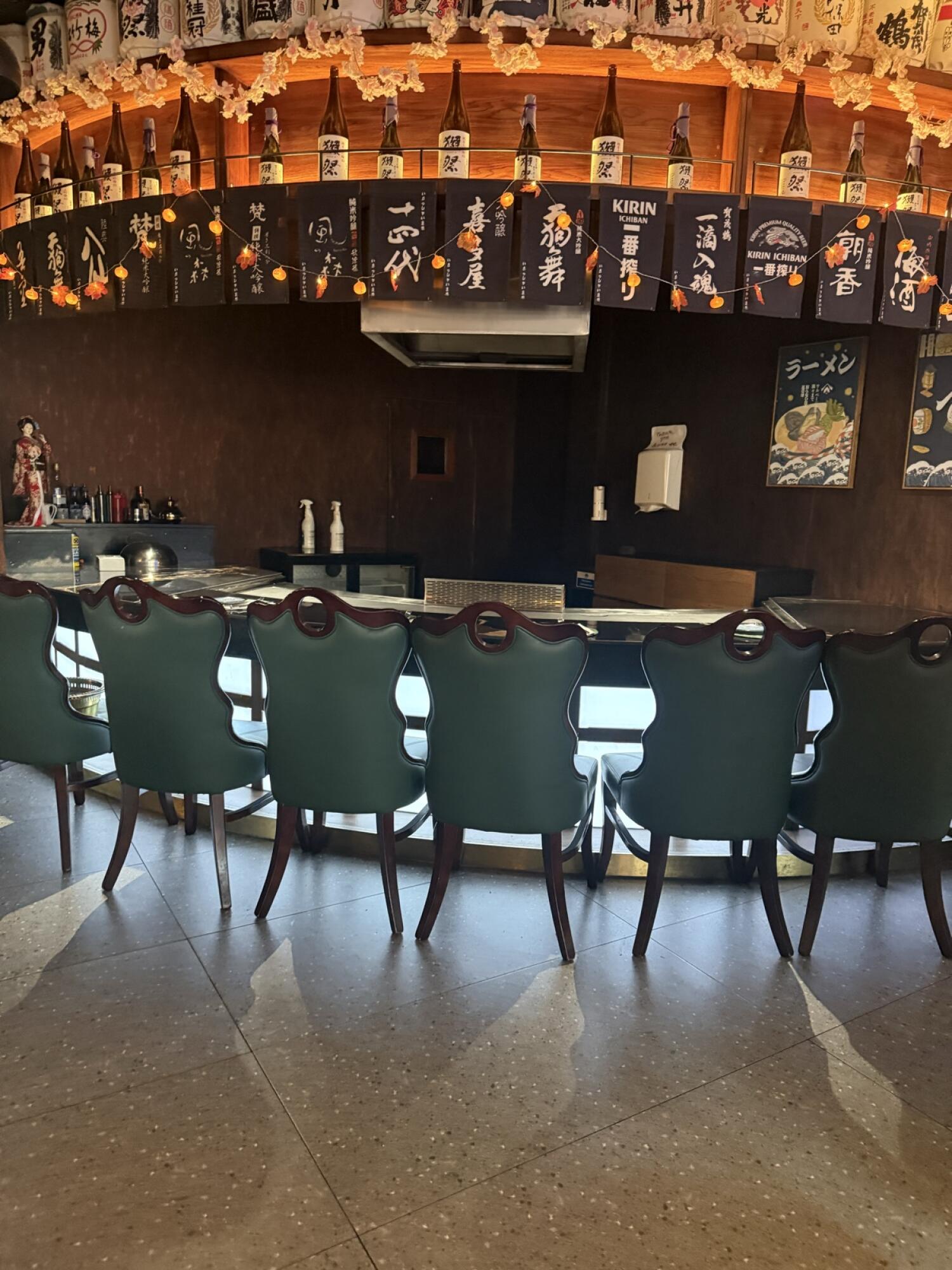 From center facing bar counter. Row of high-back chairs in front. Bottles and hanging signs displayed above counter.