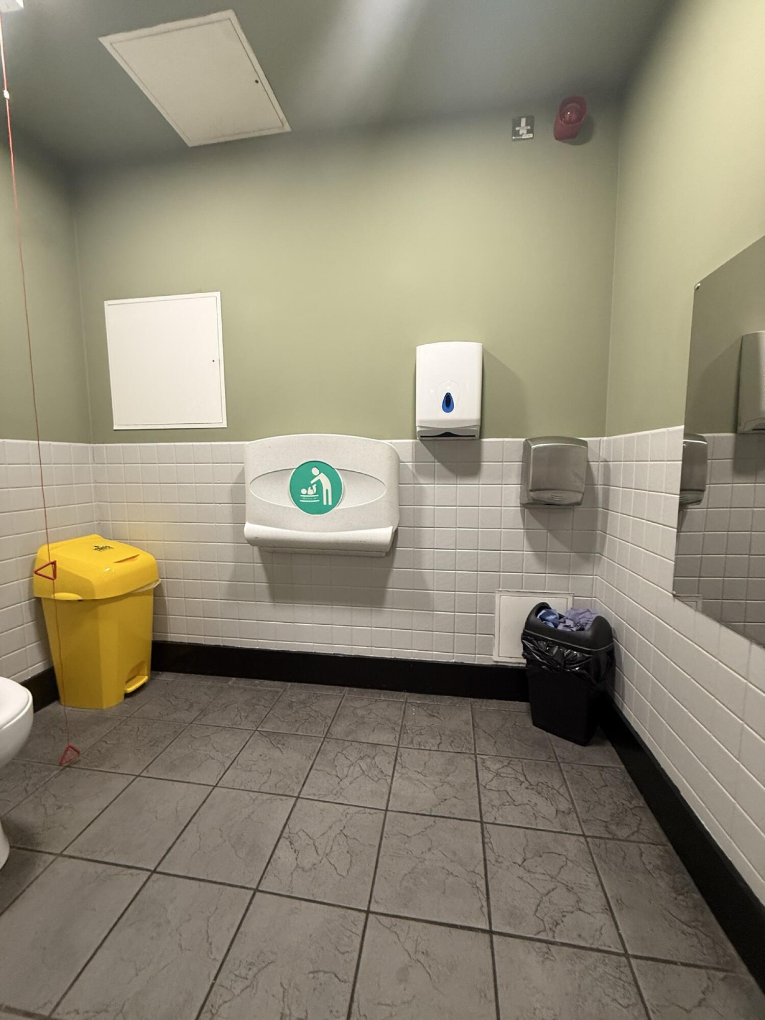 From bathroom entrance facing straight. Baby changing station in centre. Yellow bin left. Soap dispenser and bin right.