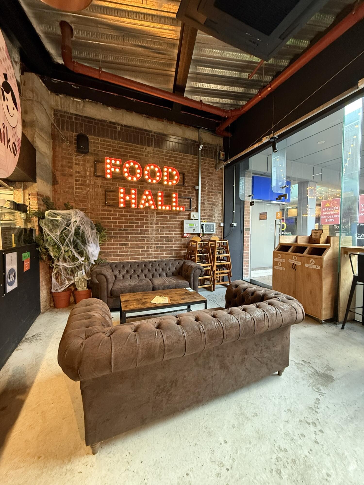From inside seating area facing entrance. Two brown sofas around low table centre. Baby high chairs stacked back right. Bin unit right.