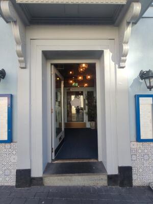 From sidewalk facing venue. White framed preceding glass door ahead. Push golden bar on right to open.