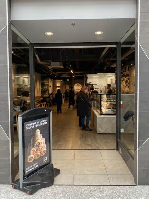 From pathway facing the venue. Entrance ahead. Framed sign on left. Glass wall on left and right side of opening. Black framed opening at back.