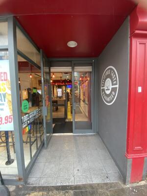 From sidewalk facing the venue. Opening ahead. Grey framed glass door at back.Pull grey metal handle from centre to open.