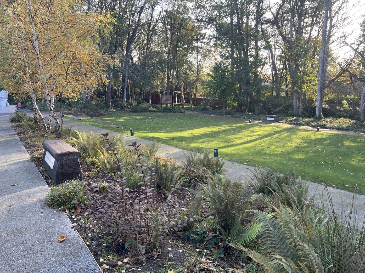 From path facing straight. Grass lawn to right with small lights along edge. Trees and shrubs border lawn, with benches along paths.