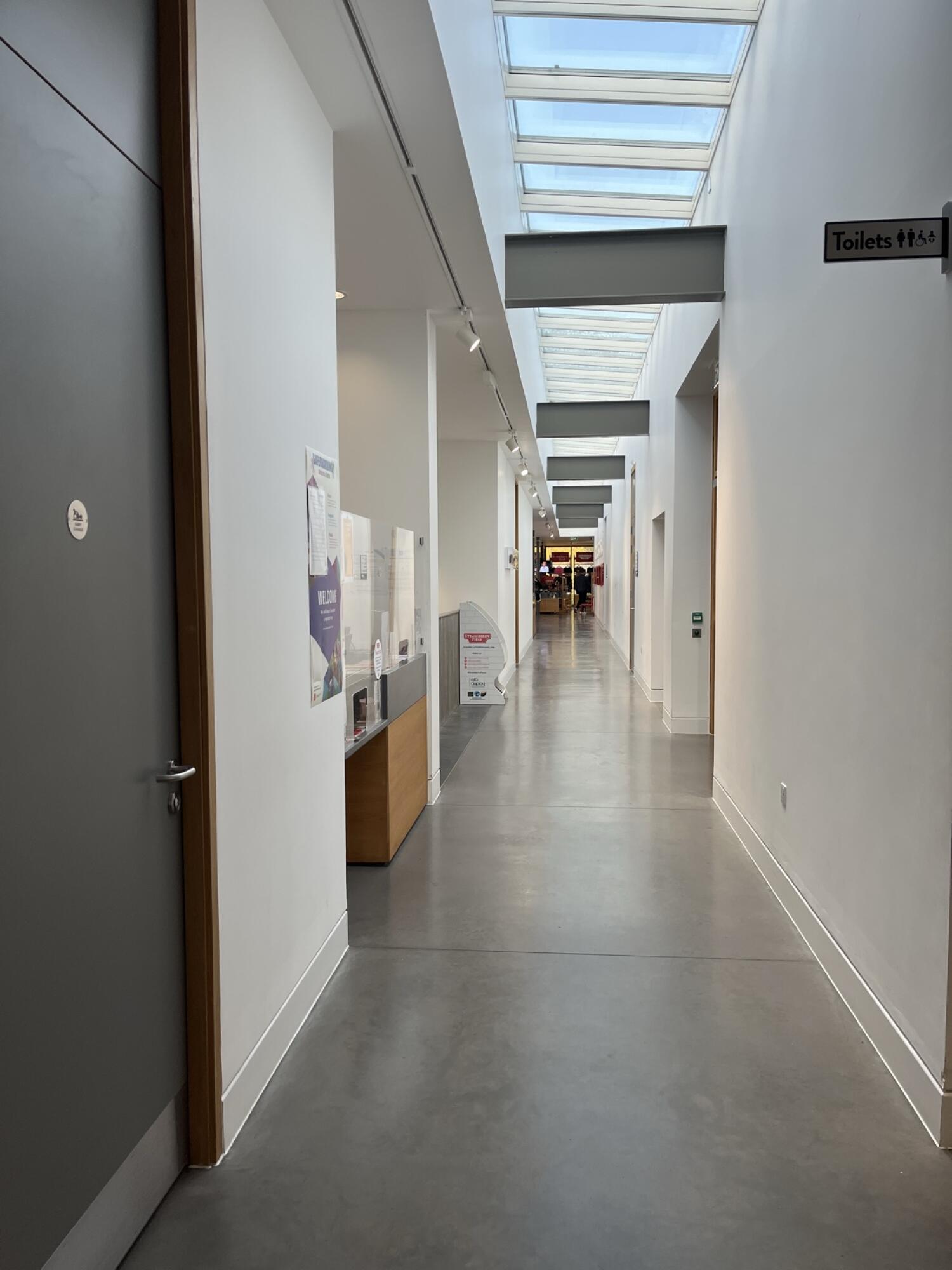From corridor facing straight. Door on left at start of corridor. Toilets sign on right ahead. Path continues to main space at end.
