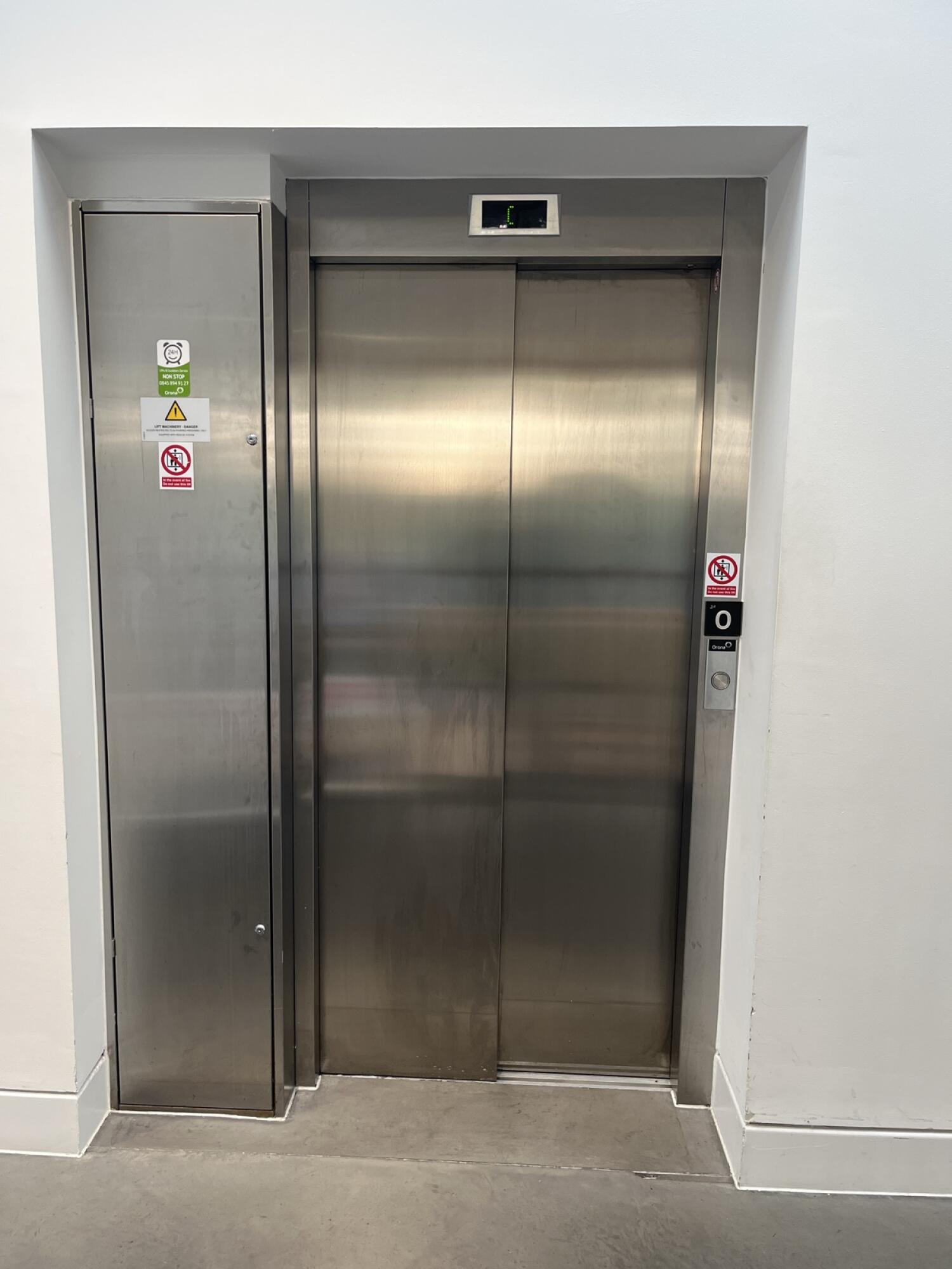 From hallway facing straight. Closed stainless steel lift doors with control panel on right. Small metal cabinet with warning signs on left.