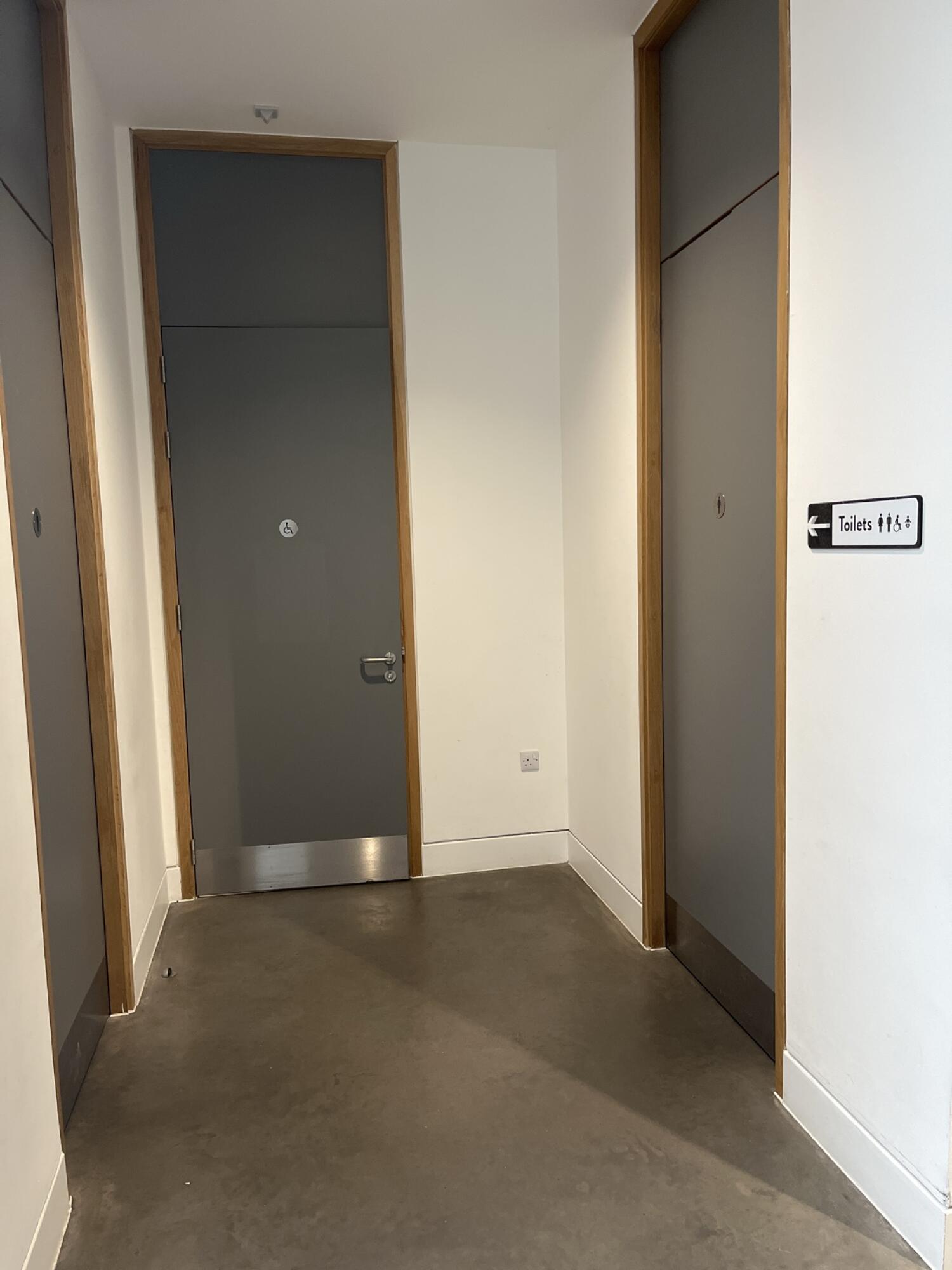 From hallway facing straight. Grey door ahead with toilet symbol. Grey door on right. Toilet sign on right wall pointing left.