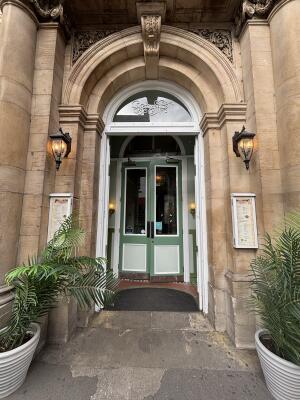 From sidewalk facing venue. White doors ahead with planters on both sides. Push from centre to open.