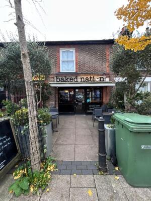 From street facing venue. Opening ahead between tree on left and bollard on right. Path behind between outdoor seating to entrance doors on back.