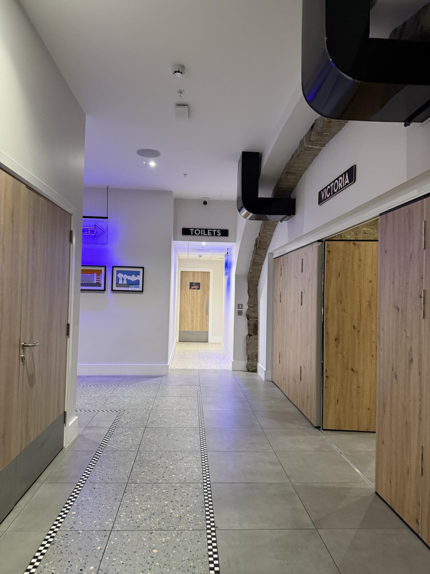 From hallway facing straight. Door to toilets at end of hall. Wooden doors along left and right. Victoria sign above right door.