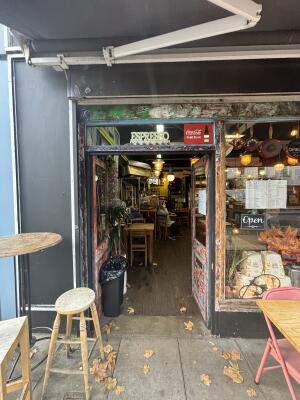 From sidewalk facing entrance. Red framed wooden glass doors ahead. Doors open from centre inwards.