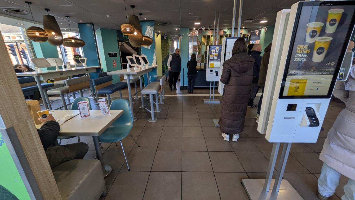 From left of kiosks facing right. Kiosk along right. Path in centre. Divider, half booths, and chairs along left, high tables with stools behind.