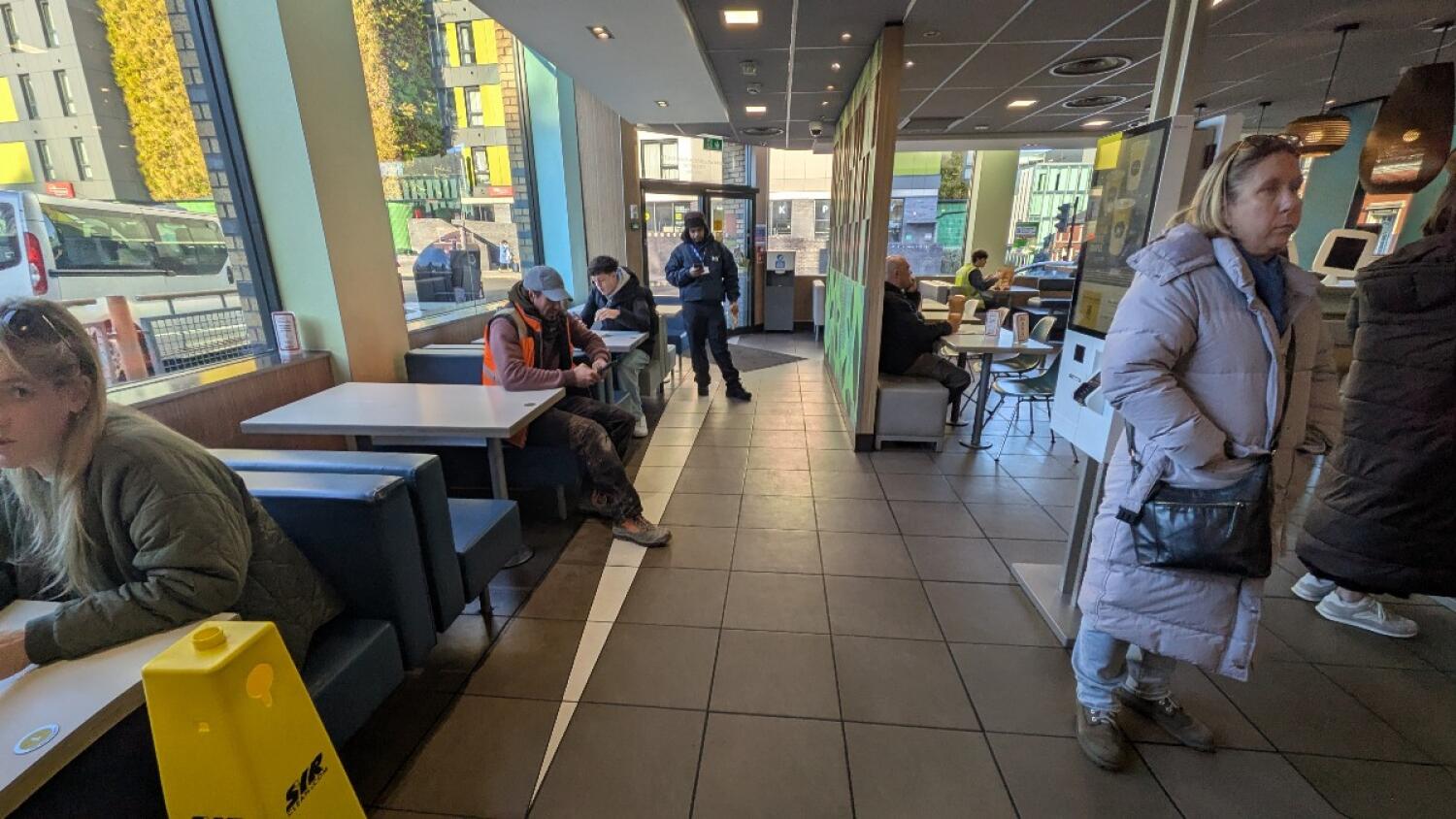 From after counter facing right. Booths along left. Path in centre leading to alternate entrance. Kiosks on right. Tables and chairs along back right.