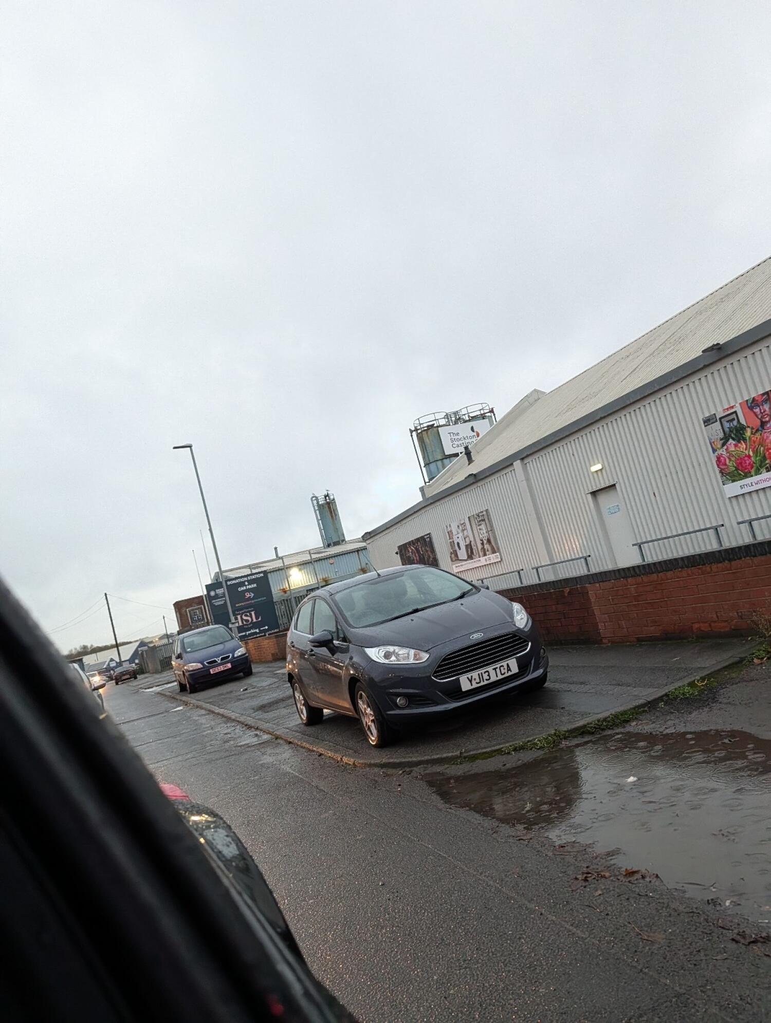 Grey car parked on pavement right. Industrial building with signage behind. Road extends ahead with more parked cars on left side.