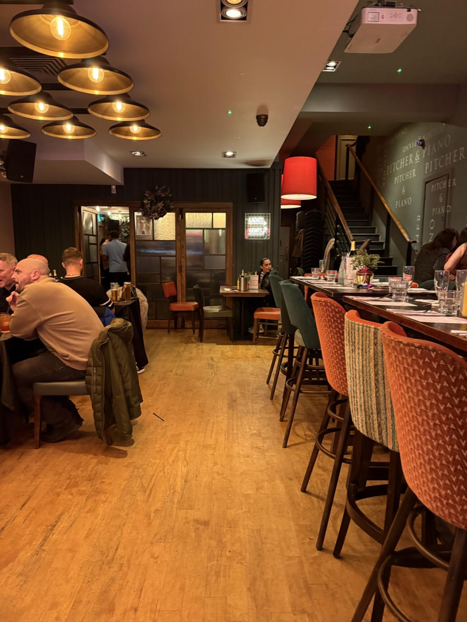 Bar counter with high stools along right. Path in centre leading to doorway at back. Tables and chairs along left, staircase up at back right.